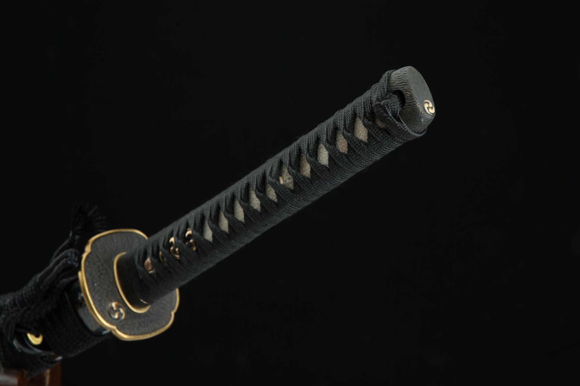Close-up of a samurai sword handle featuring a kashira with a distinctive swirl design, black cord wrap, and brass tsuba.