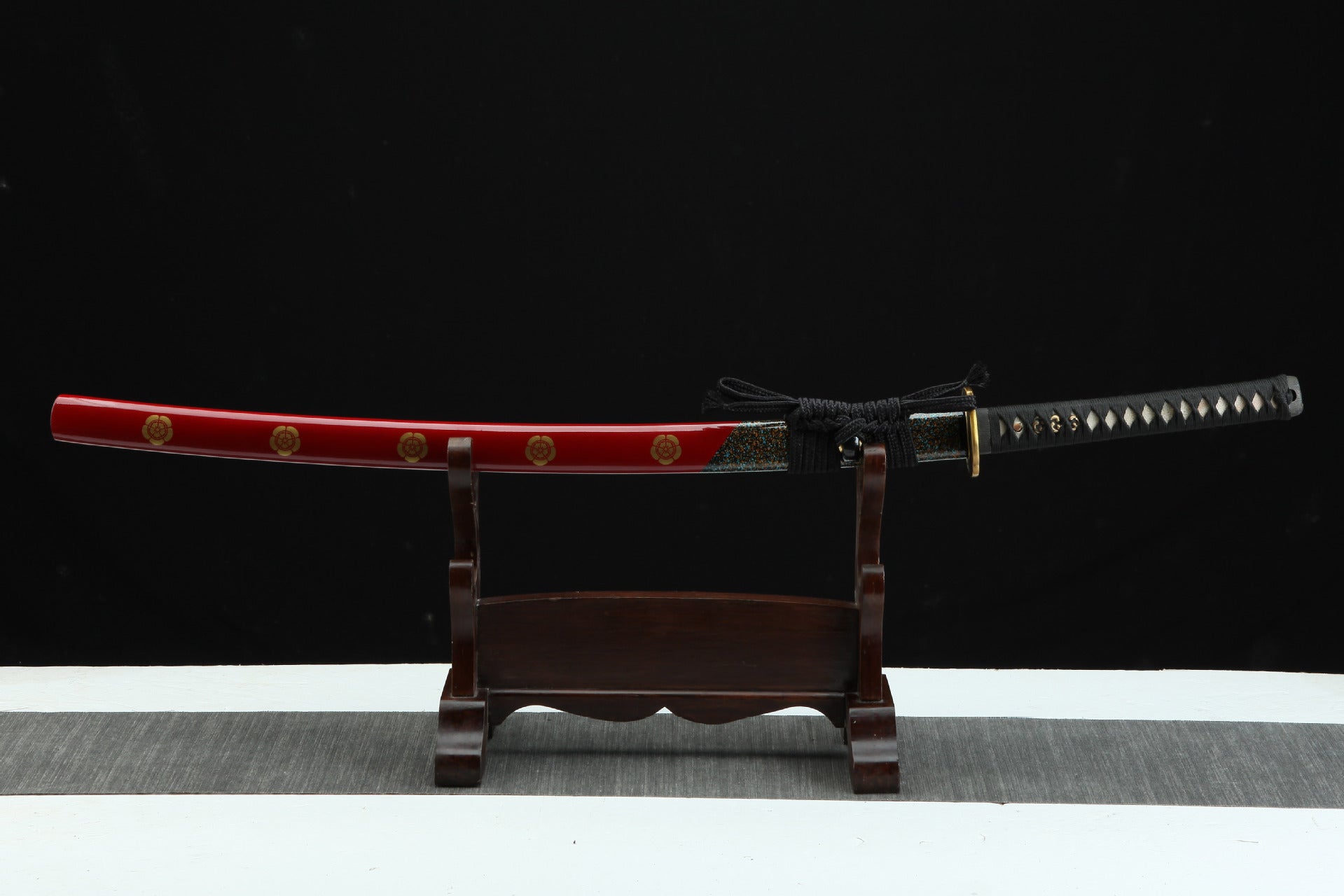 Full view of a samurai sword with a red saya featuring gold flower patterns, displayed on a wooden stand, showcasing the tsuka and tsuba.