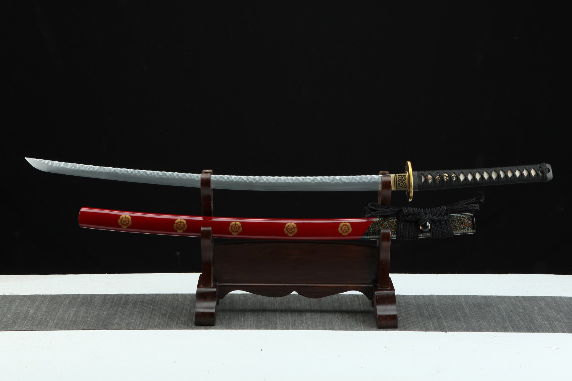 Handcrafted Damascus steel samurai sword with a red lacquered scabbard, brass tsuba, and black handle wrap. Displayed on a wooden stand.