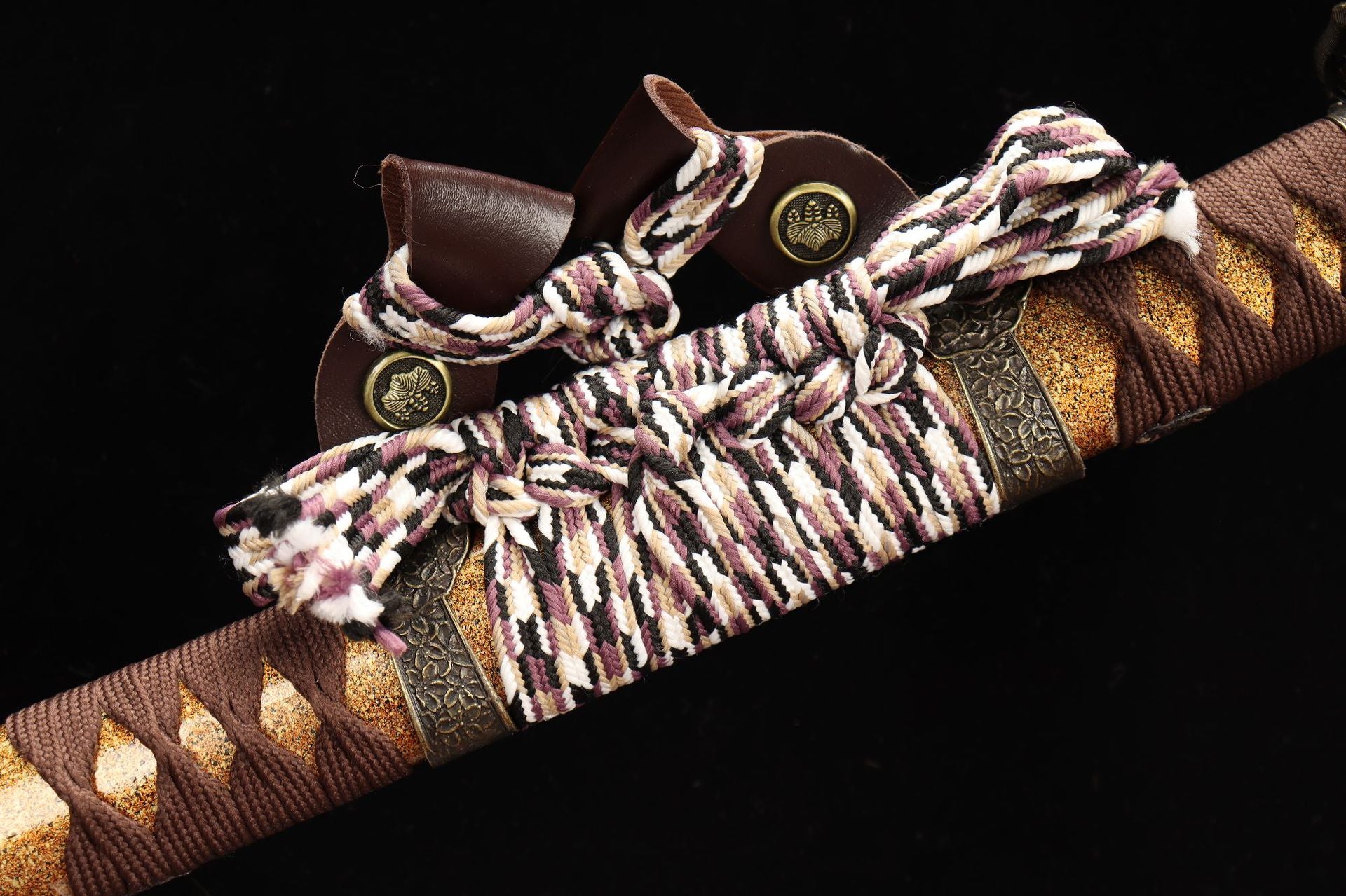 Close-up of the Nobunaga Tachi sword's sageo cord, intricately braided in purple, white, and black, attached to a golden decorative scabbard with fine metal fittings.